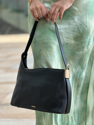 Paired Oriana Gold-Lock Bag in black, held by its strap in both hands, with a gold-tone side detail, over a green satin skirt