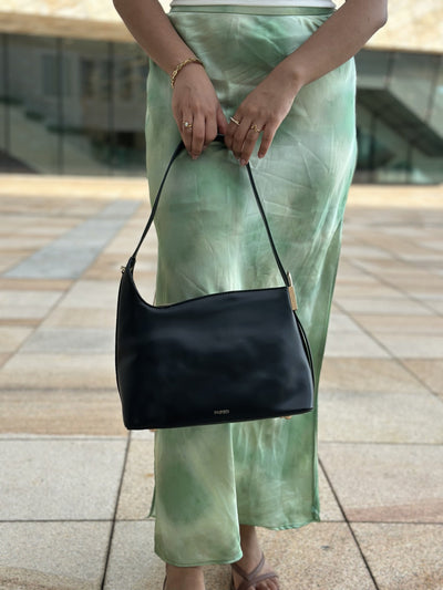 Paired Oriana Gold-Lock Bag in black, held by its strap in front of a green satin skirt, showing its soft shape