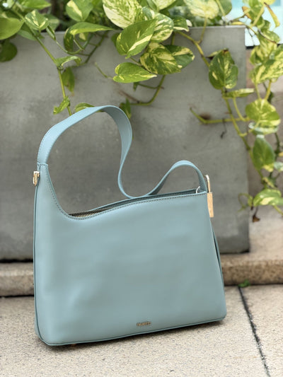 Paired Oriana Gold-Lock Bag in soft blue, a relaxed shoulder bag with a curved strap and gold-tone detail on a terrace