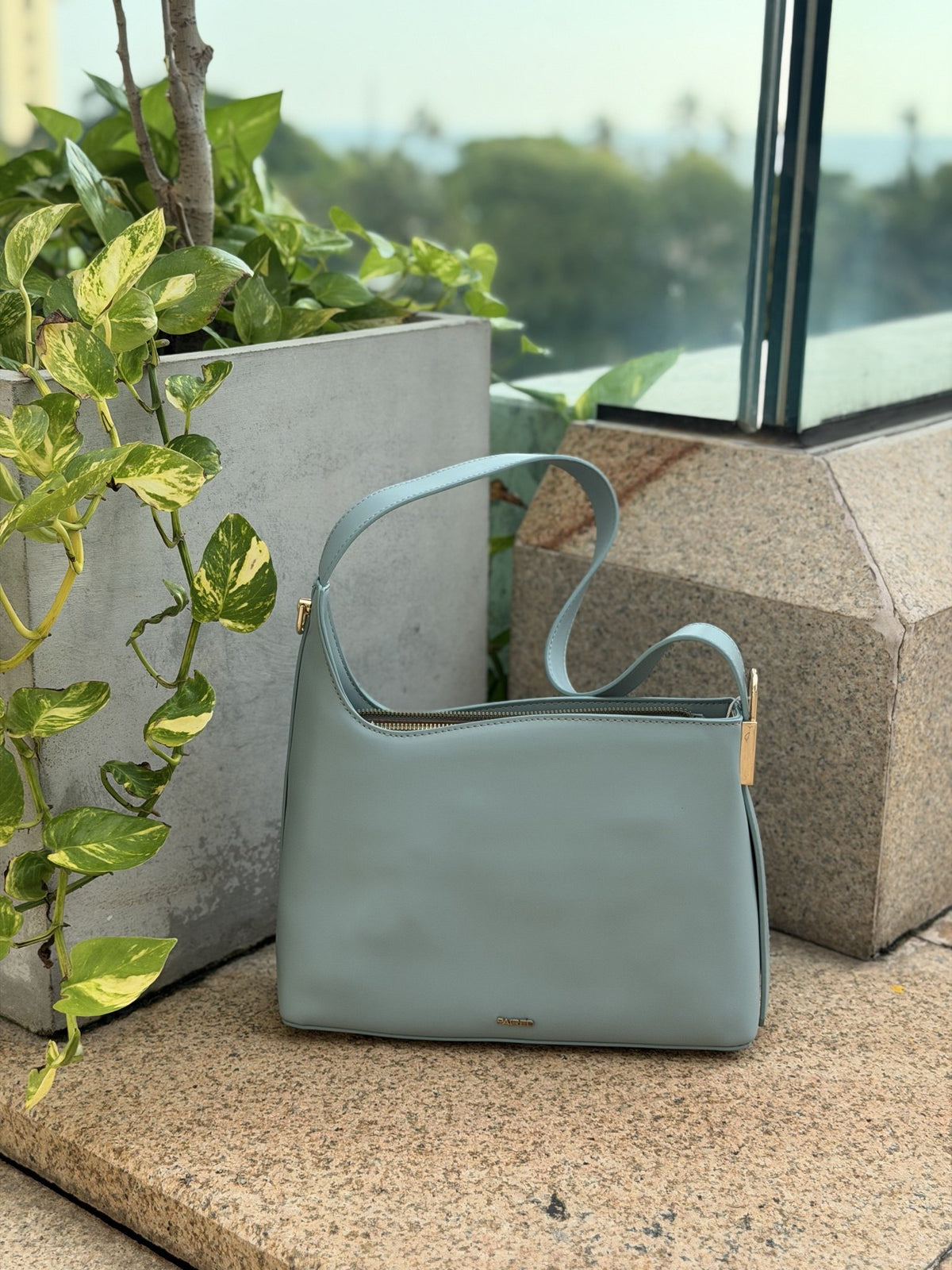 Paired Oriana Gold-Lock Bag in soft blue, a relaxed shoulder bag with a curved strap, standing on a stone ledge with greenery