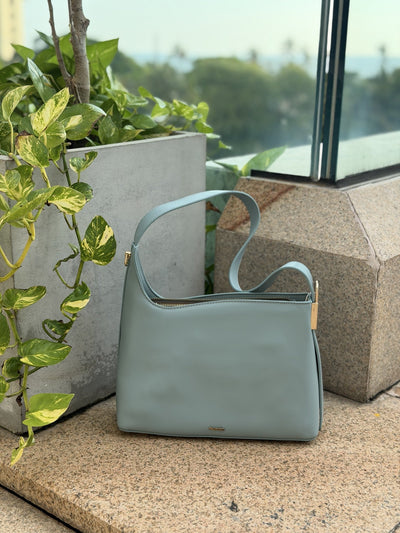 Paired Oriana Gold-Lock Bag in soft blue, a relaxed shoulder bag with a curved strap, standing on a stone ledge with greenery