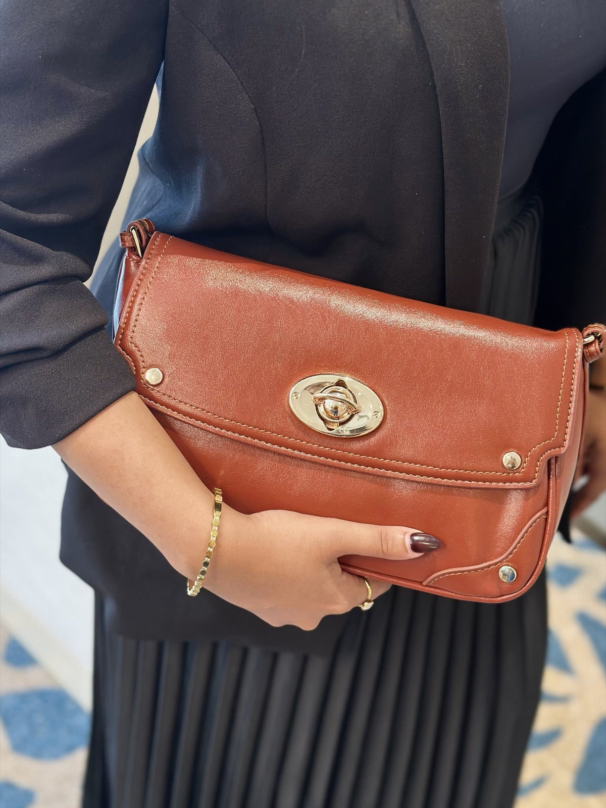 Paired Riviera Lock Bag in tan brown, carried in the hand like a clutch, with a gold-tone oval turn-lock and corner studs