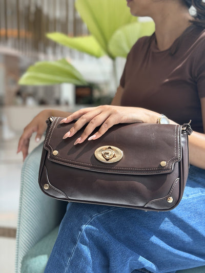 Paired Riviera Lock Bag in coffee brown, with a gold-tone oval turn-lock, resting on the lap of a woman in blue jeans