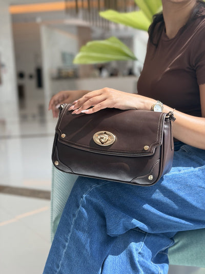 Paired Riviera Lock Bag in coffee brown, with a gold-tone oval turn-lock and studded corners, held on the lap with jeans