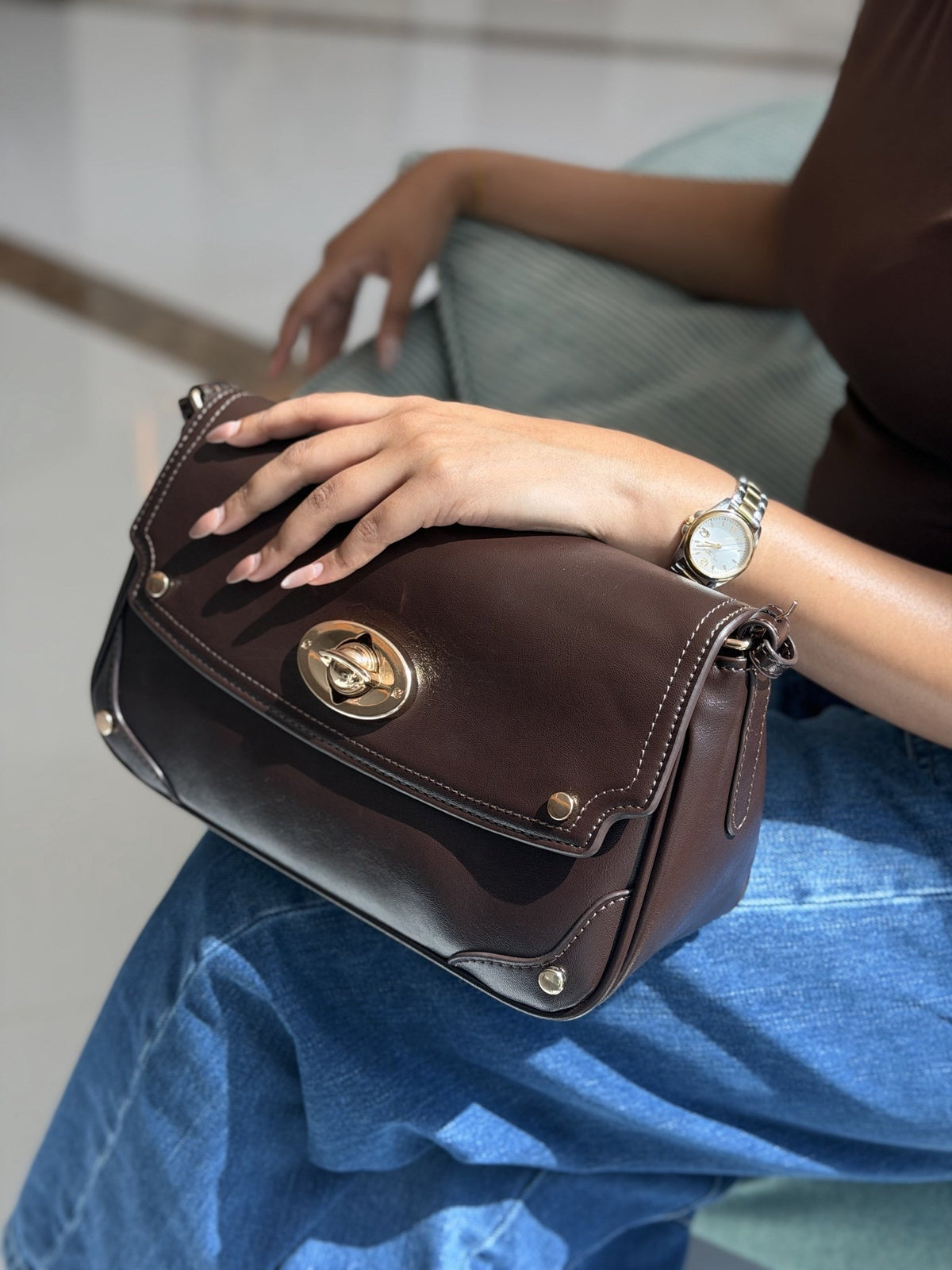 Paired Riviera Lock Bag in coffee brown, with a gold-tone oval turn-lock, held on the lap with a hand resting on top