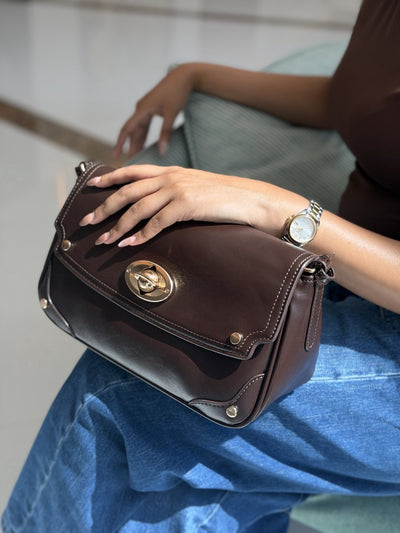 Paired Riviera Lock Bag in coffee brown, with a gold-tone oval turn-lock, held on the lap with a hand resting on top