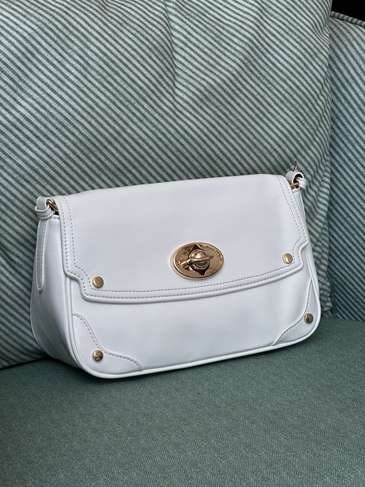 Paired Riviera Lock Bag in white, with a flap, gold-tone oval turn-lock and studded corners, resting on a pale green armchair