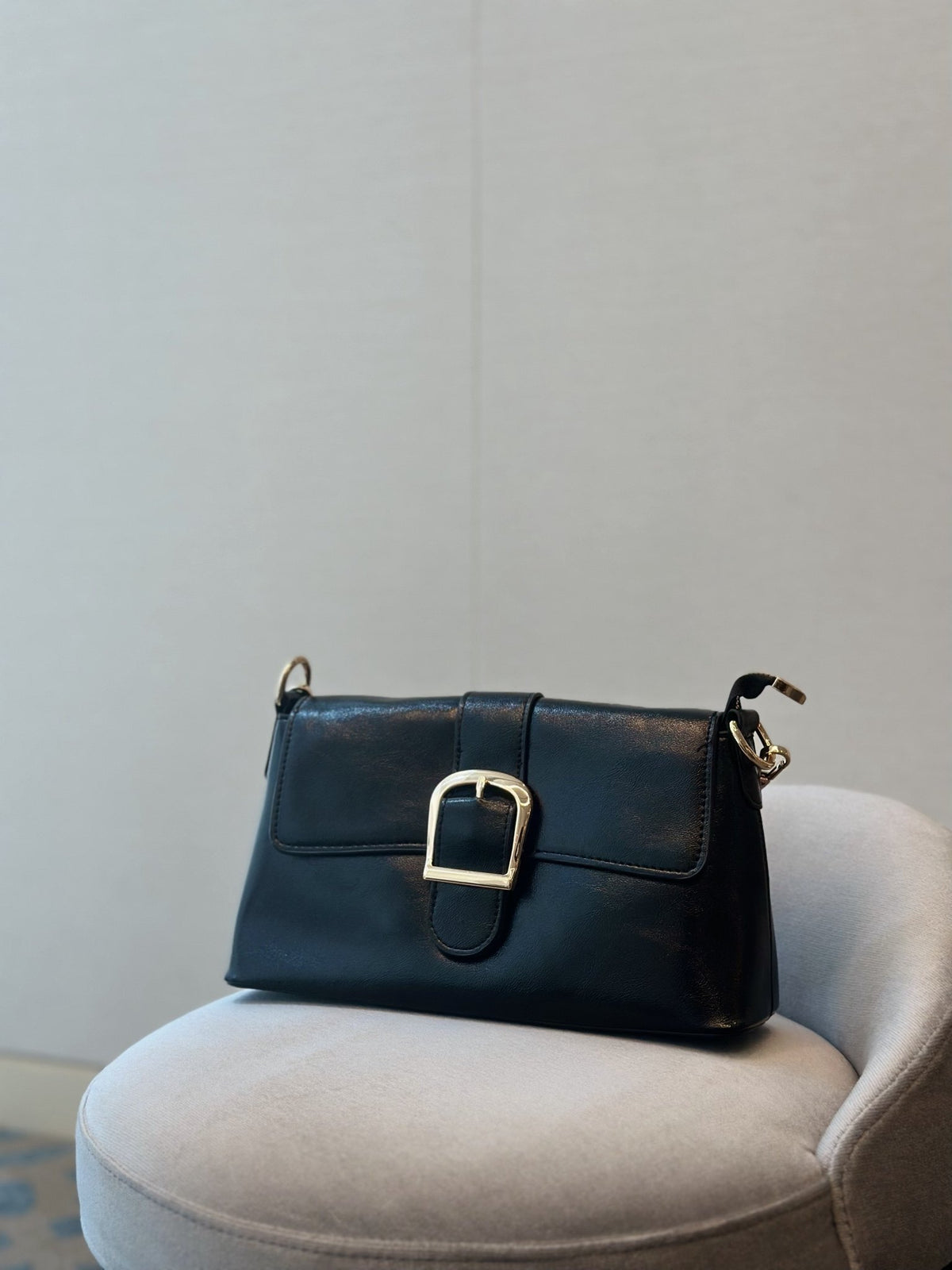 Paired Valora Buckle Sling Bag in black, with a buckled front flap, sitting on a round grey seat