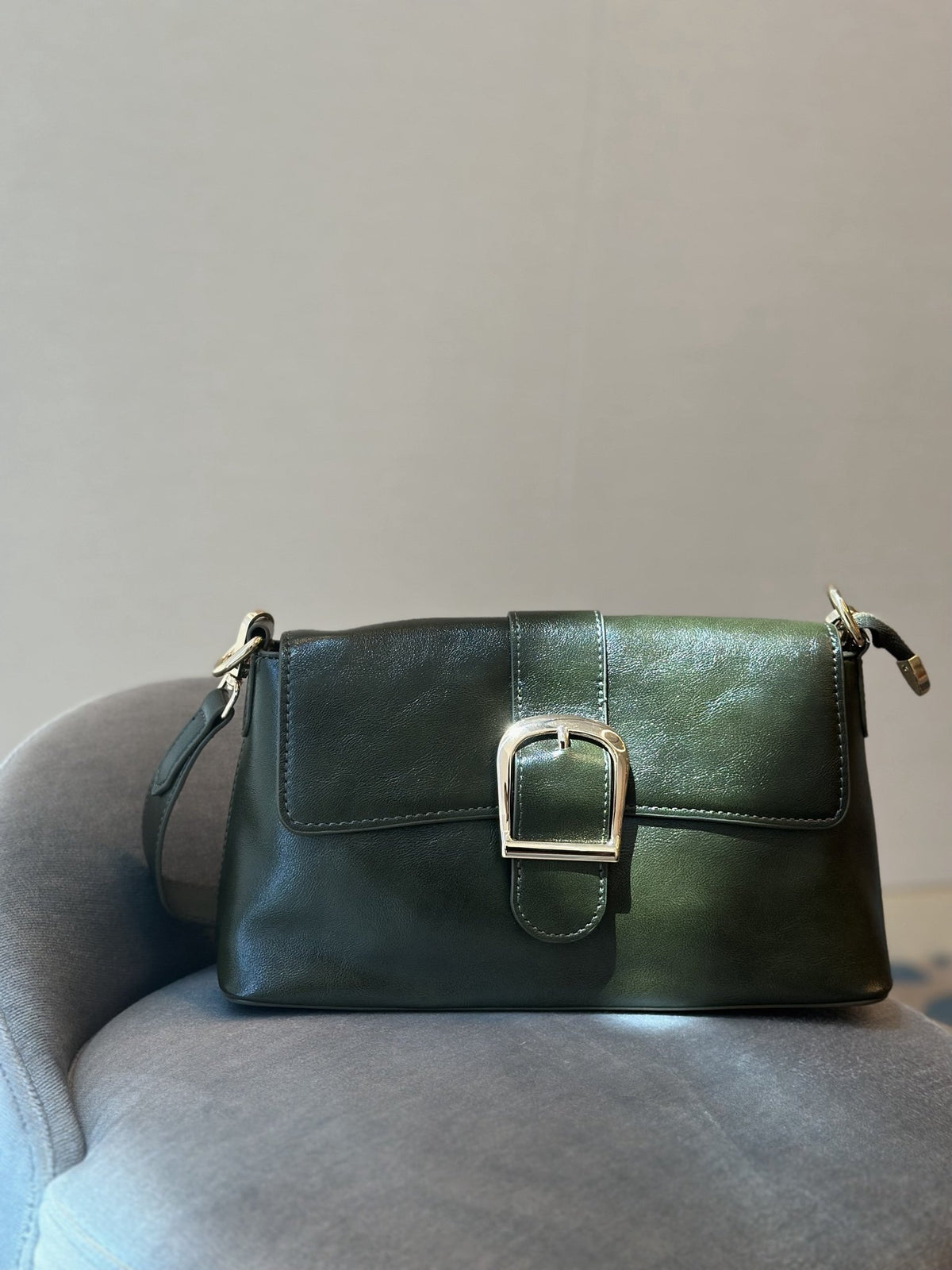 Paired Valora Buckle Sling Bag in deep green, with a buckled front flap, resting on a grey chair