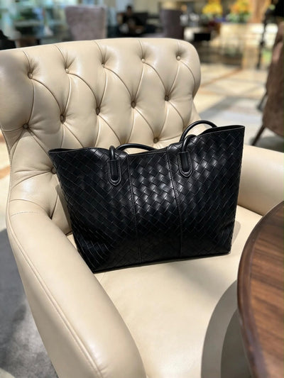Paired Virelle Woven Bag in black, resting on a cream armchair, showing the woven texture and two rolled handles