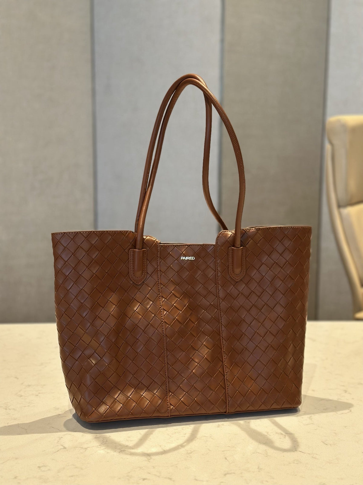 Paired Virelle Woven Bag in tan brown, an open-top tote with two rolled handles and woven texture on a marble table