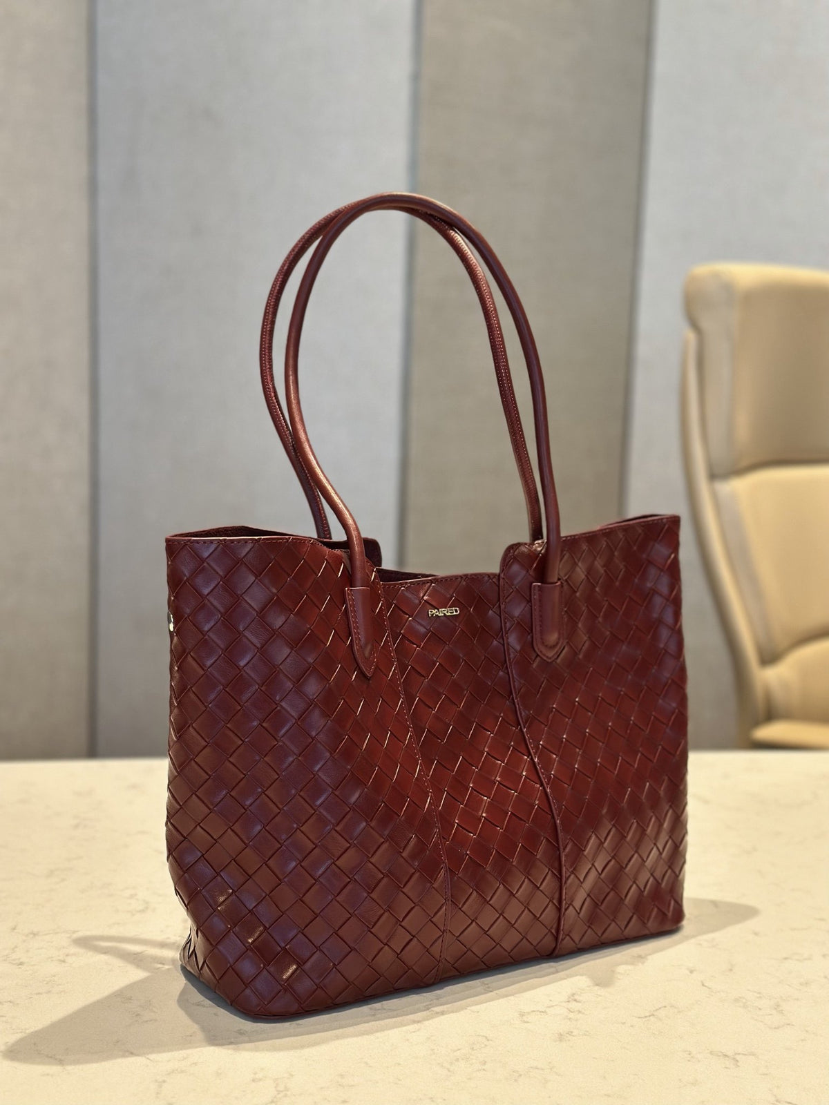 Paired Virelle Woven Bag in maroon, an open-top tote with two rolled handles, shown at a slight angle on a marble table
